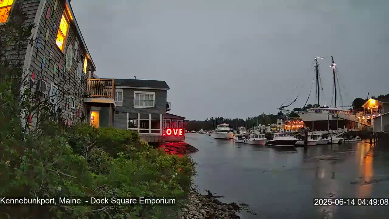 A harbor scene at dusk shows a waterfront building with a red "LOVE" sign, boats docked along a calm waterway, and a large sailing ship moored nearby under an overcast sky.