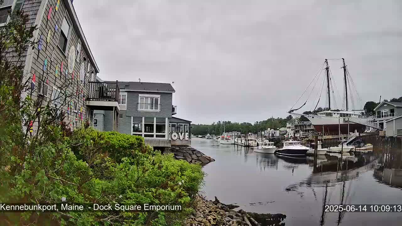 A harbor scene on an overcast day shows several boats docked, with buildings lining the waterway, one of which is decorated with colorful nautical flags.