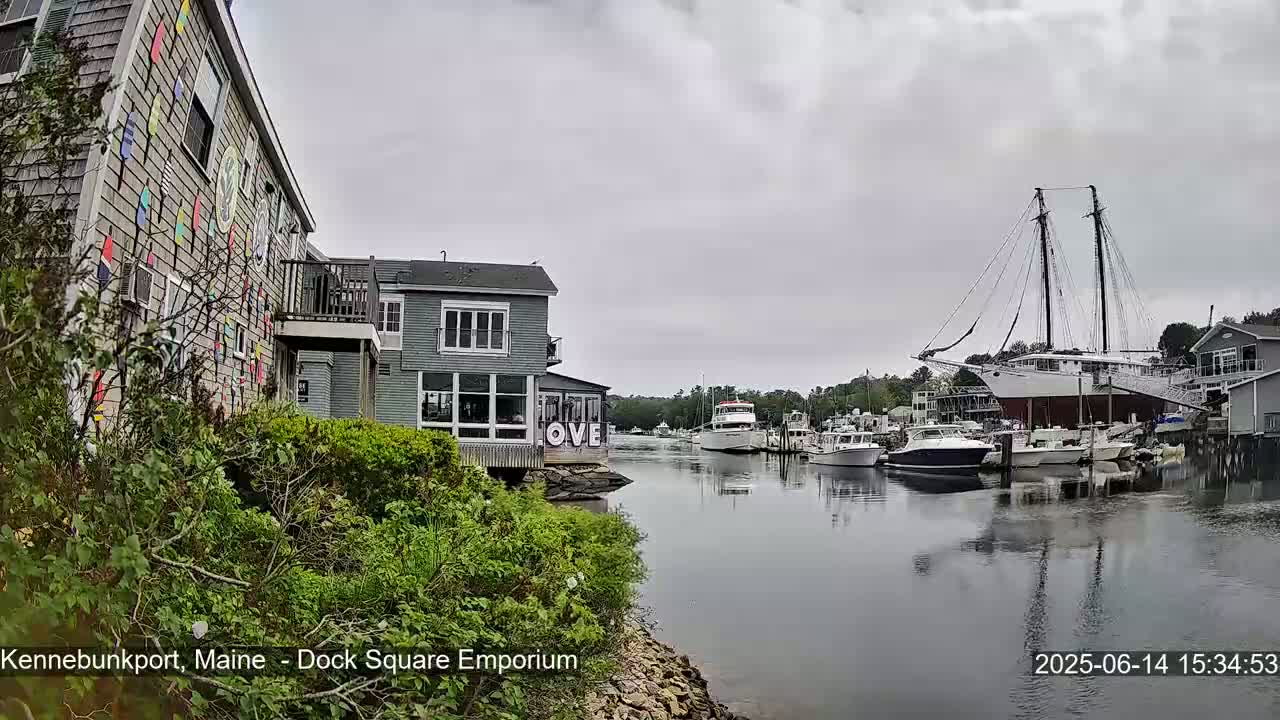 A harbor scene on an overcast day shows several boats docked, with buildings lining the waterway, one of which is decorated with colorful nautical flags.