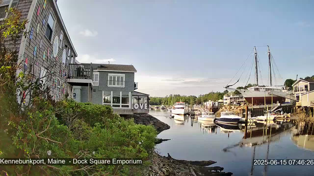 A calm harbor scene on a sunny day features several boats, including a large sailboat docked near waterfront buildings, one of which displays the word "LOVE."