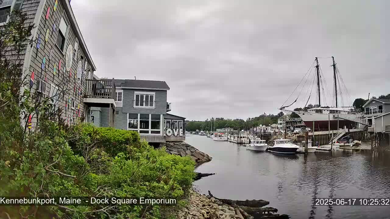 A gray-shingled building decorated with colorful pennants sits beside a calm waterway where several boats are moored, under a cloudy sky.
