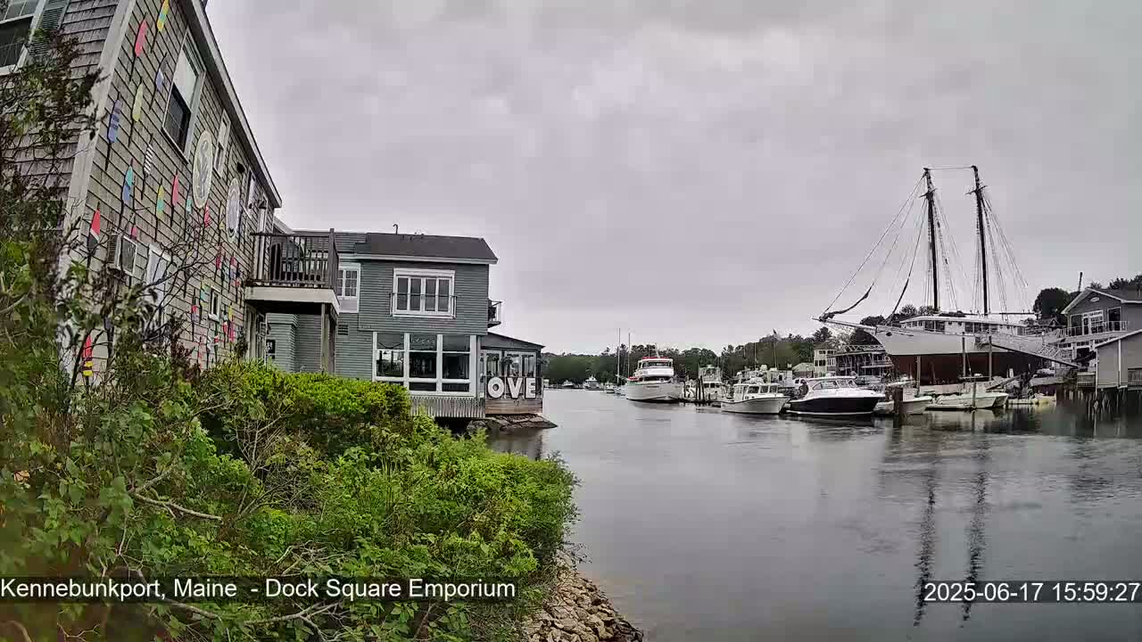A waterfront scene on an overcast day shows a canal with several boats, including a large sailboat, and buildings with colorful decorations along the shore.