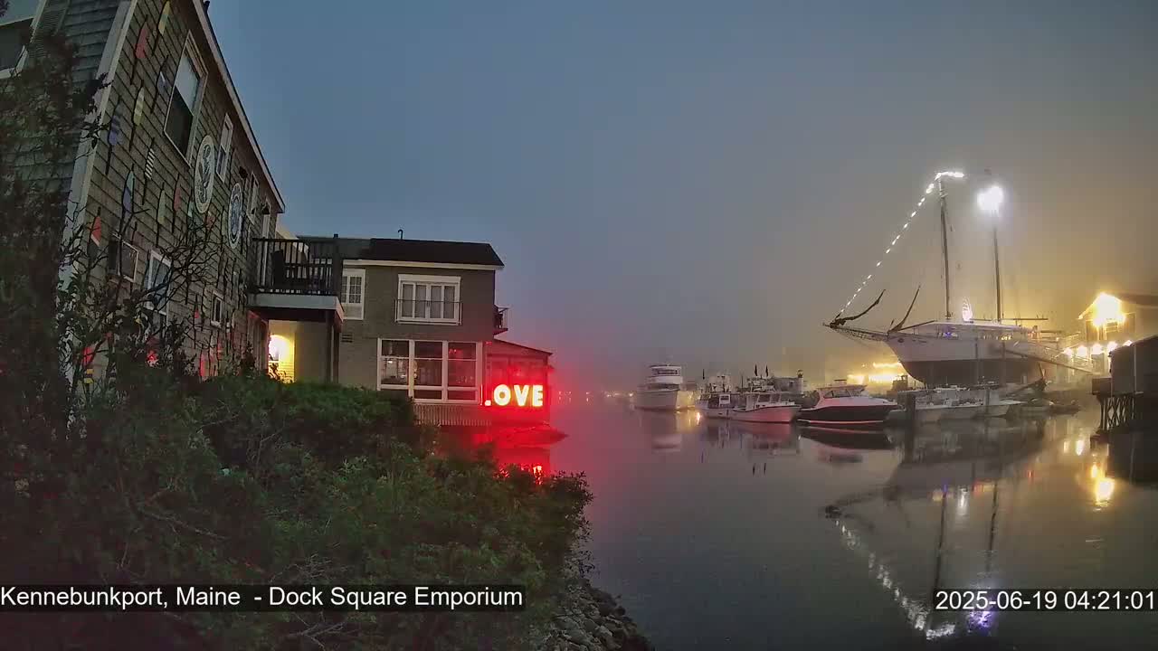 A foggy harbor at night, featuring a large sailboat, several smaller boats, and a building with a "LOVE" sign illuminated.