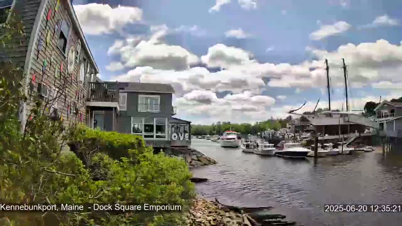 A partly cloudy day shows a waterfront scene with colorful buildings, docked boats, and lush green vegetation.