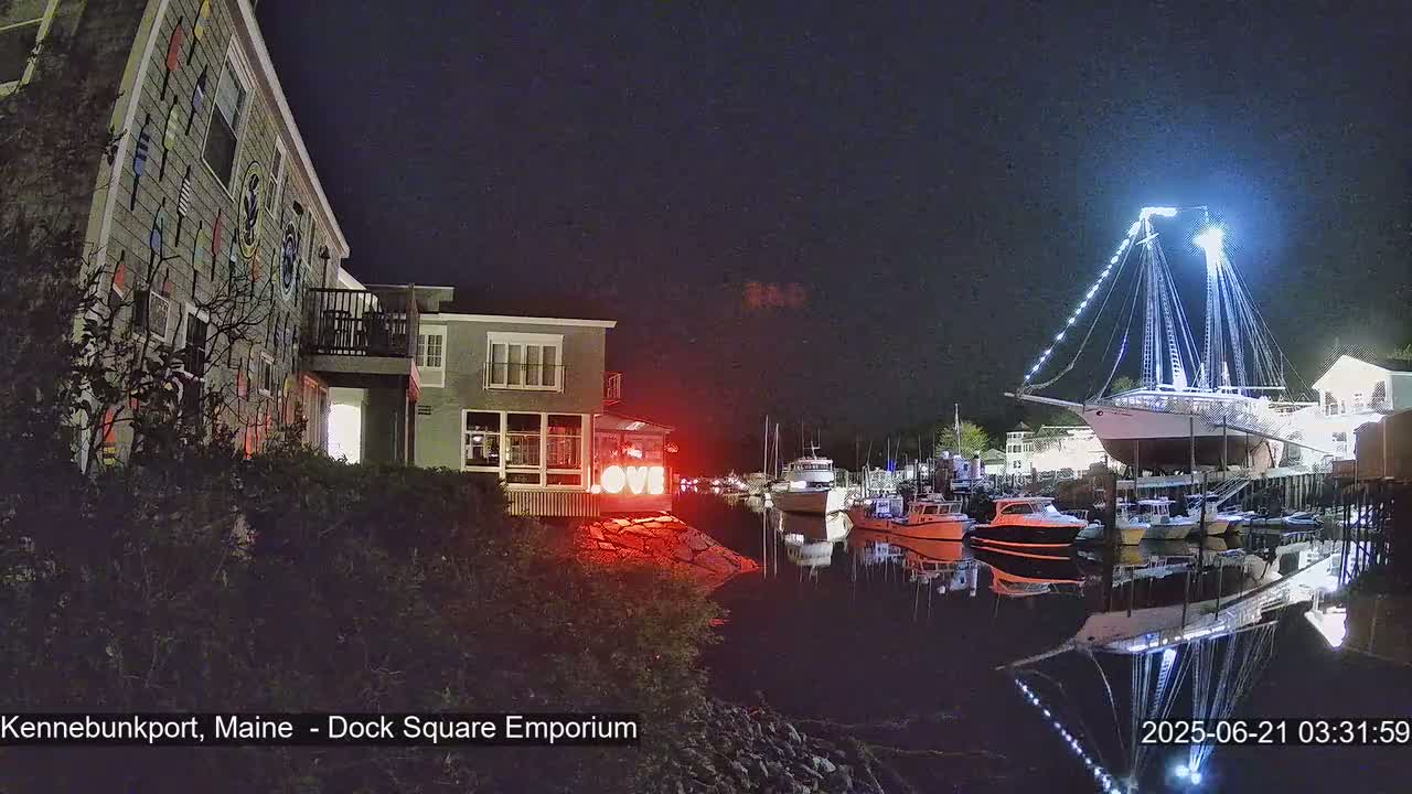 A brightly lit sailboat is docked at night next to a waterfront building decorated with nautical flags, with several smaller boats in the harbor.
