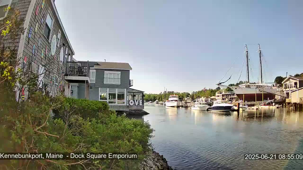 A calm harbor scene on a sunny day shows a colorful building, other buildings along the waterfront, several boats docked, and a large sailboat at the center.