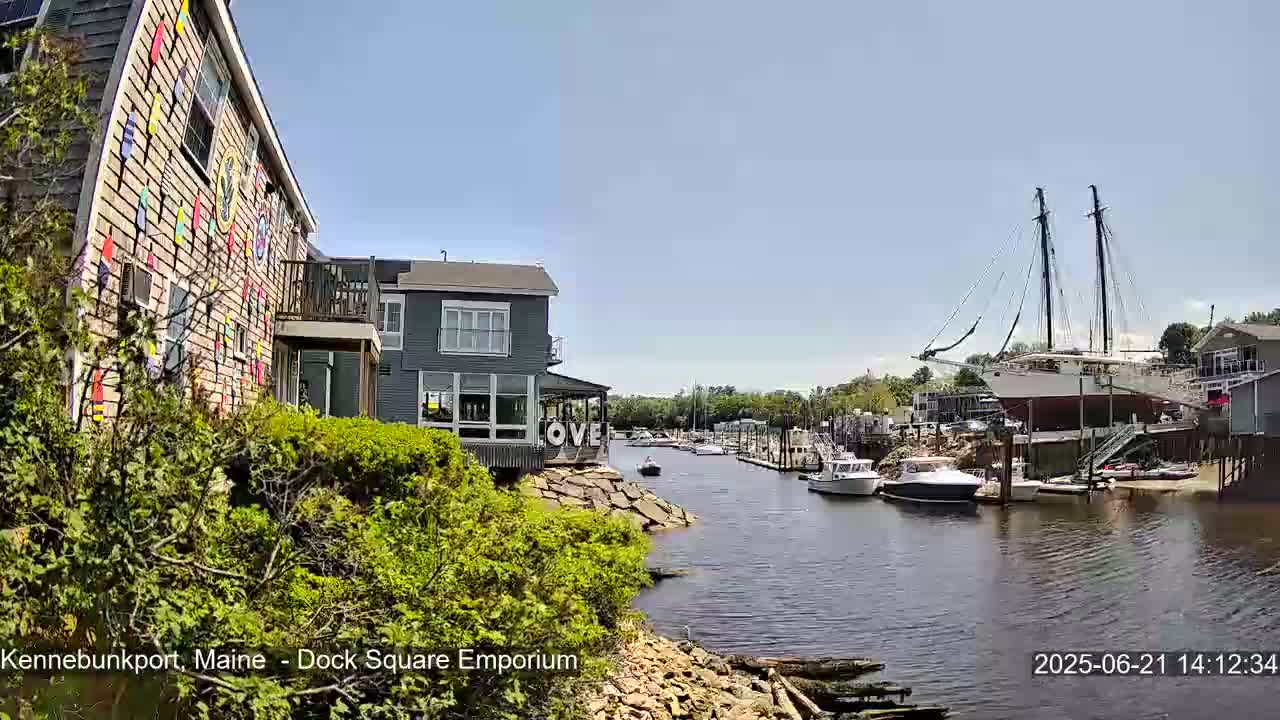 A waterfront scene on a sunny day shows a canal with moored boats, buildings along the shore, and a large sailboat on a cradle.