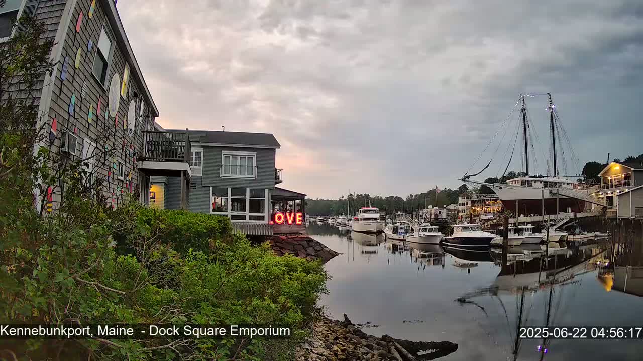 A harbor scene at dusk shows a calm body of water with several boats moored, including a large sailing ship, and buildings along the shore, one with a "LOVE" sign.