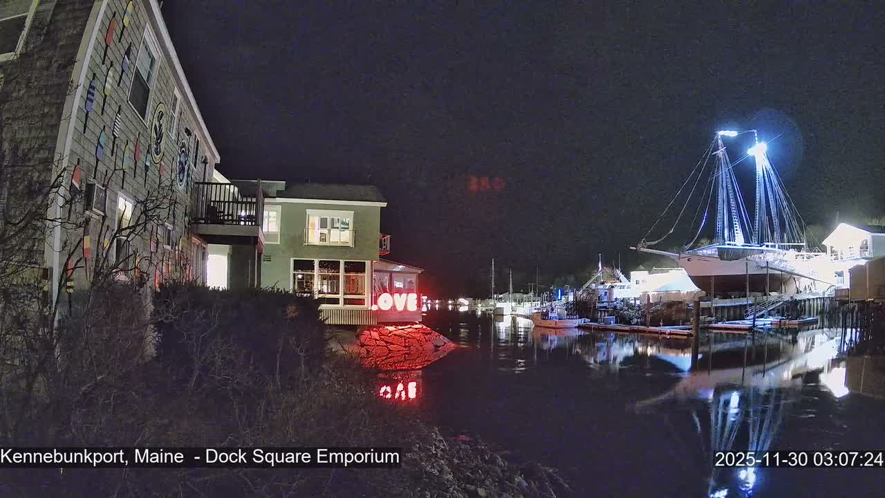 The image displays a clear night scene of a waterfront bustling with brightly lit buildings, including one with a "LOVE" sign reflecting in the dark water, and a prominent, illuminated sailboat docked by the shore.