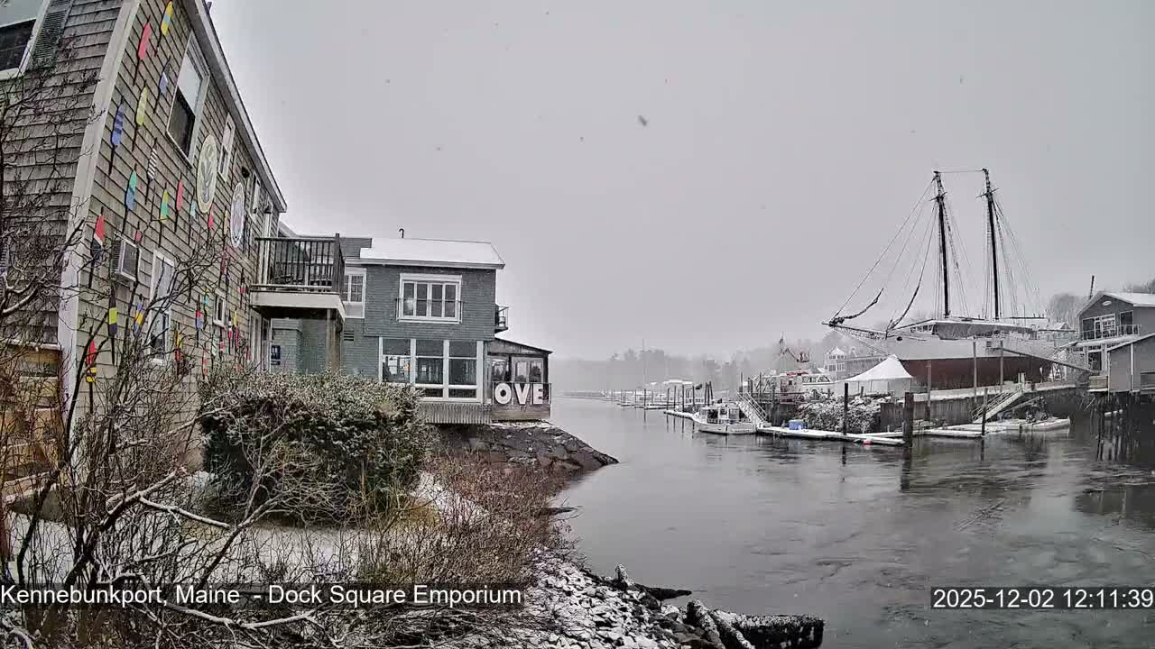 A light snowfall blankets a bustling winter harbor scene with colorful buildings, numerous docked boats, and a large schooner dry-docked on the right, all beneath a grey, overcast sky.