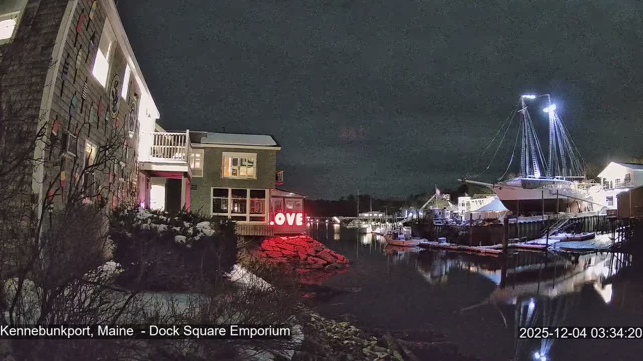 A snowy, cloudy winter night reveals a vibrant harbor scene with illuminated buildings, including one displaying a red "LOVE" sign, and a large, brightly lit schooner reflected in the calm water.