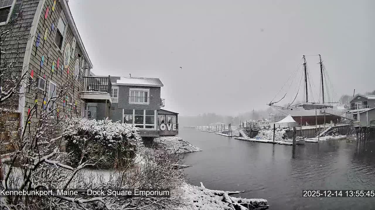 A snowy, cloudy winter night reveals a vibrant harbor scene with illuminated buildings, including one displaying a red "LOVE" sign, and a large, brightly lit schooner reflected in the calm water.