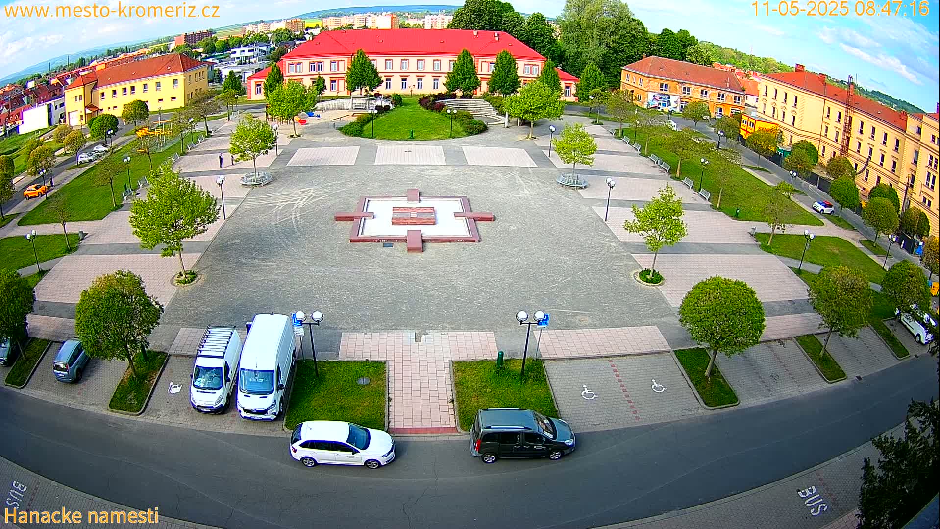 A town square with a central fountain, surrounded by buildings, green spaces, and parked cars, under a partly cloudy sky.