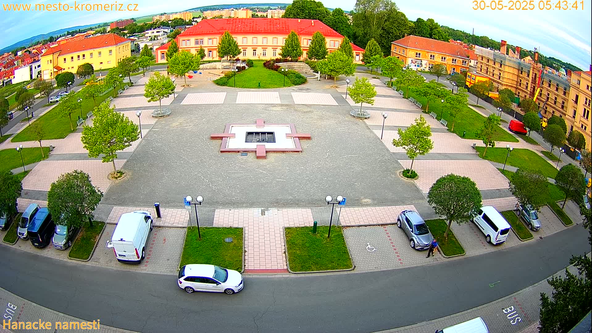An aerial view of a town square with a central fountain, surrounded by parked cars, green spaces, and buildings, under a partly cloudy sky.
