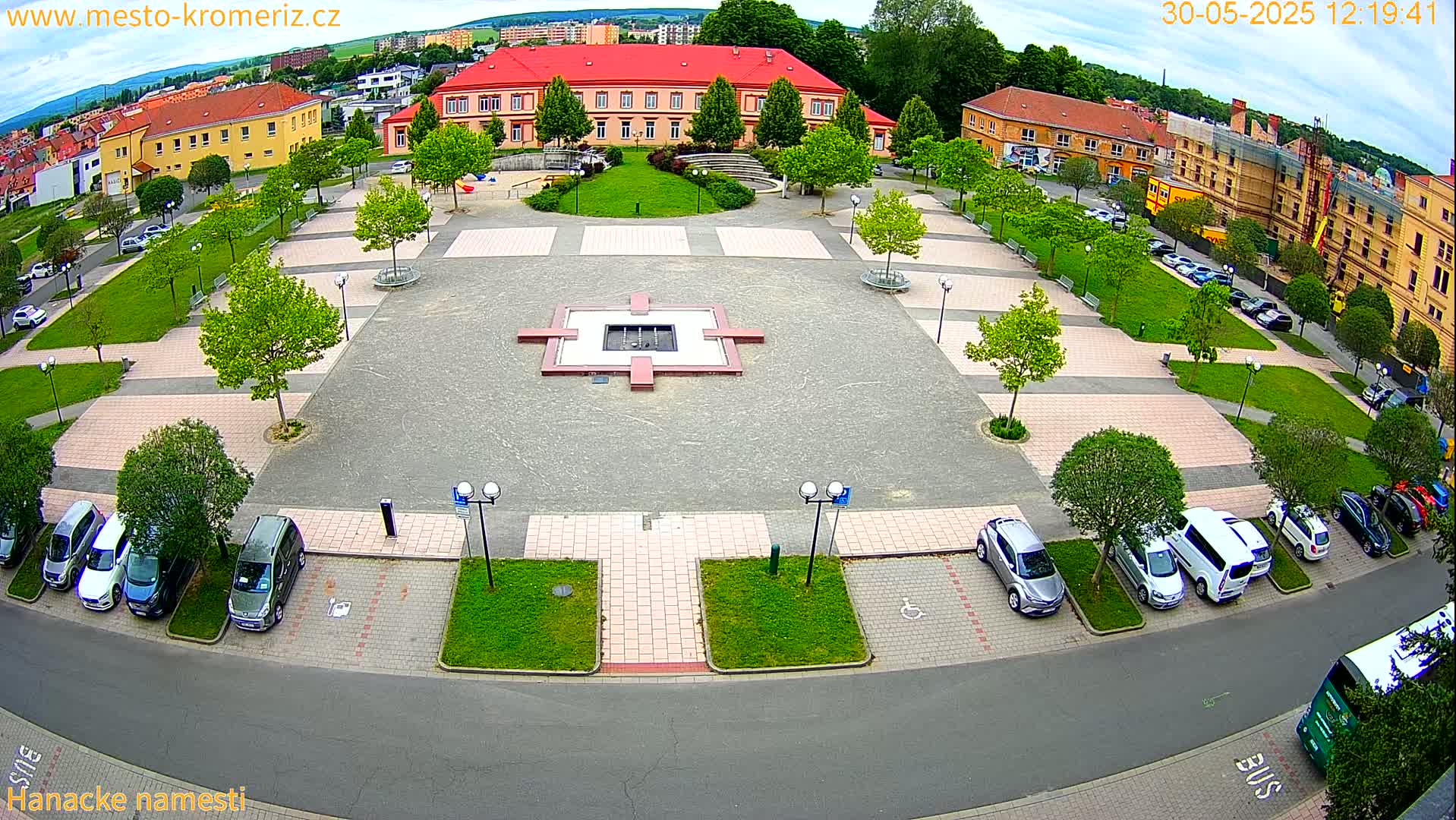 An aerial view of a town square with a central fountain, surrounded by green spaces, parked cars, and buildings, under partly cloudy skies.