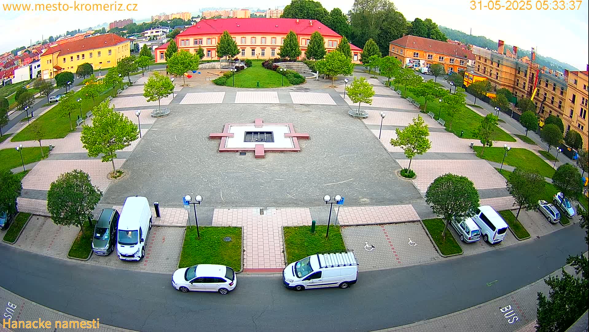 An aerial view of a town square with a central fountain, surrounded by parked cars and buildings under a partly cloudy sky.