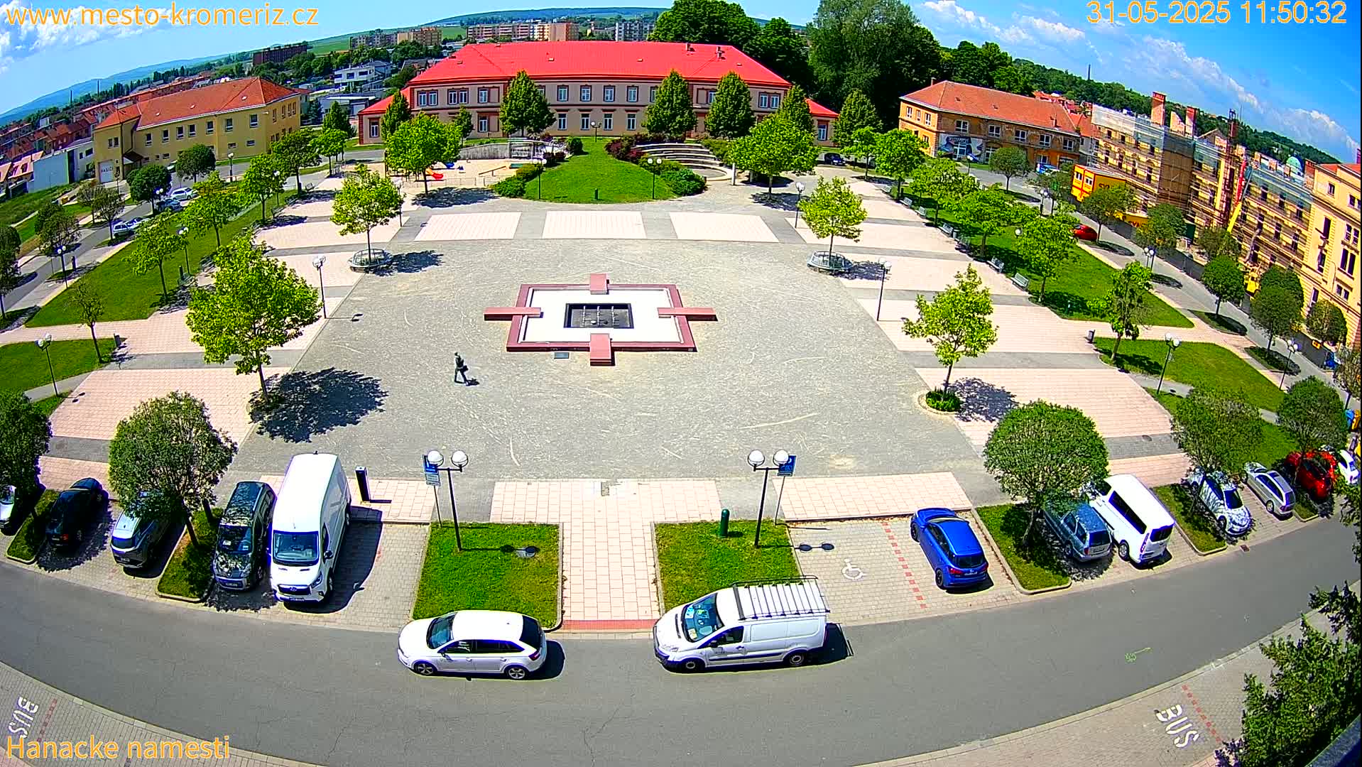 A sunny aerial view of a town square with a central fountain, surrounded by buildings, green spaces, and parked cars.