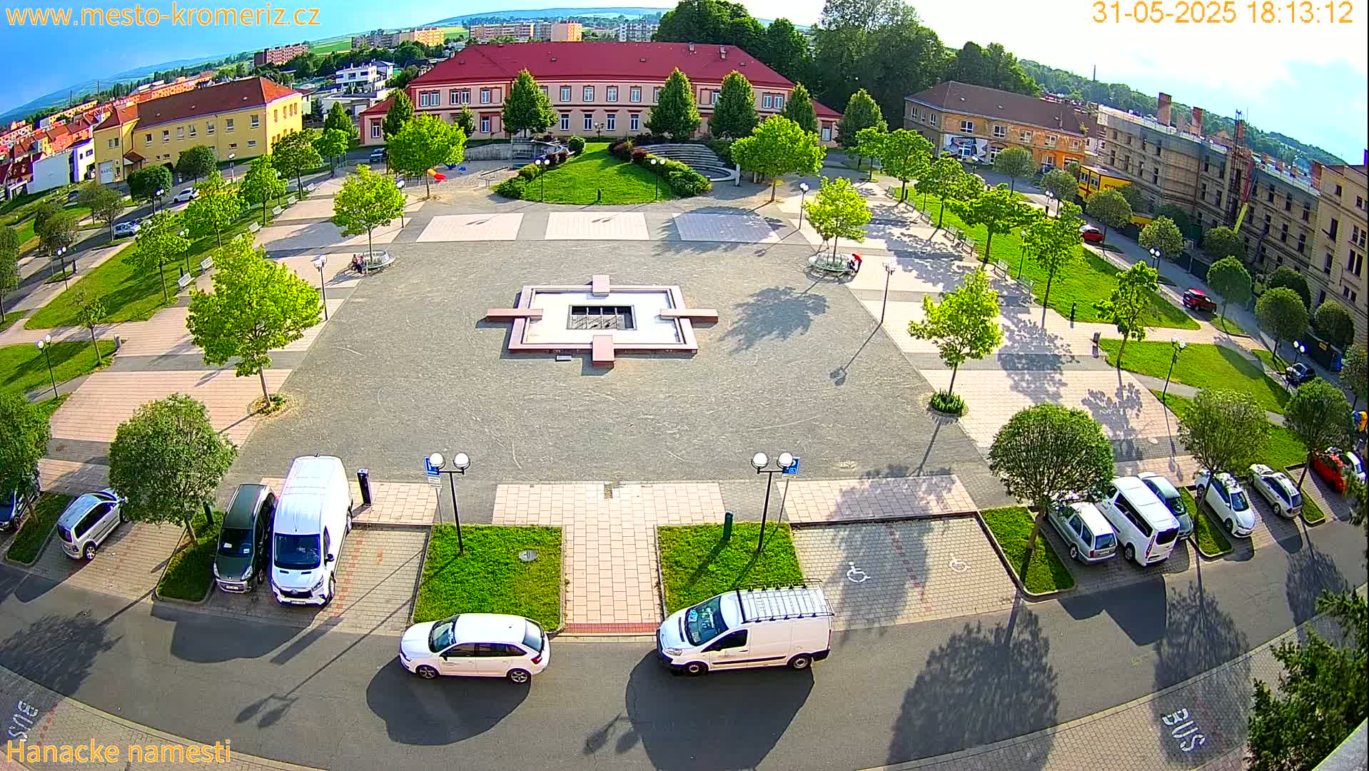 A sunny aerial view of a town square with a fountain, surrounded by buildings, trees, and parked cars.