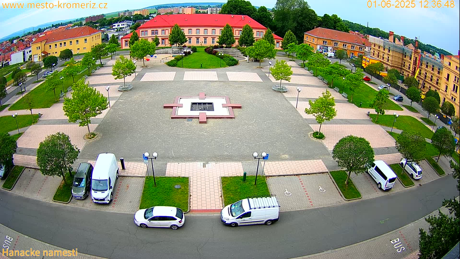 An aerial view of a town square with a fountain, surrounded by buildings, parked cars, and green spaces under partly cloudy skies.