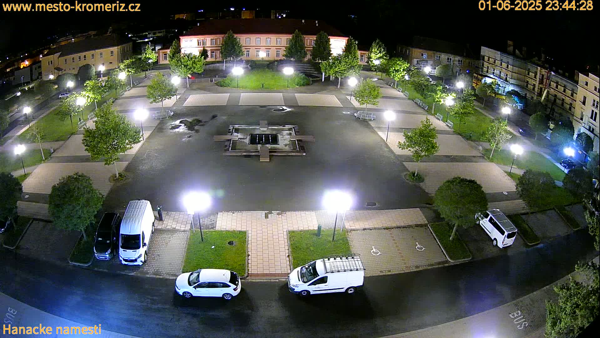 A nighttime aerial view of a town square with a fountain at its center, surrounded by parked cars, streetlights, and small green spaces, under slightly wet conditions.