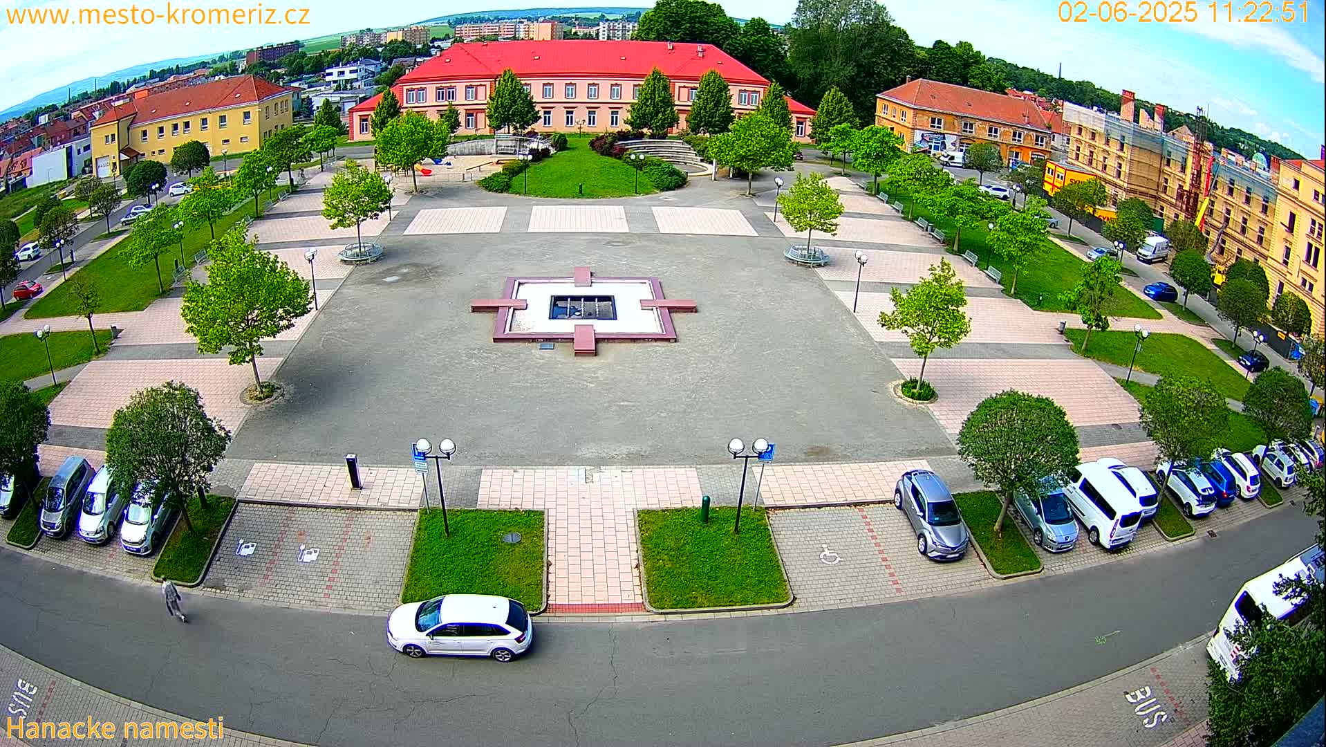 An aerial view of a town square with a central fountain, surrounded by buildings, green spaces, and parked cars under a partly cloudy sky.