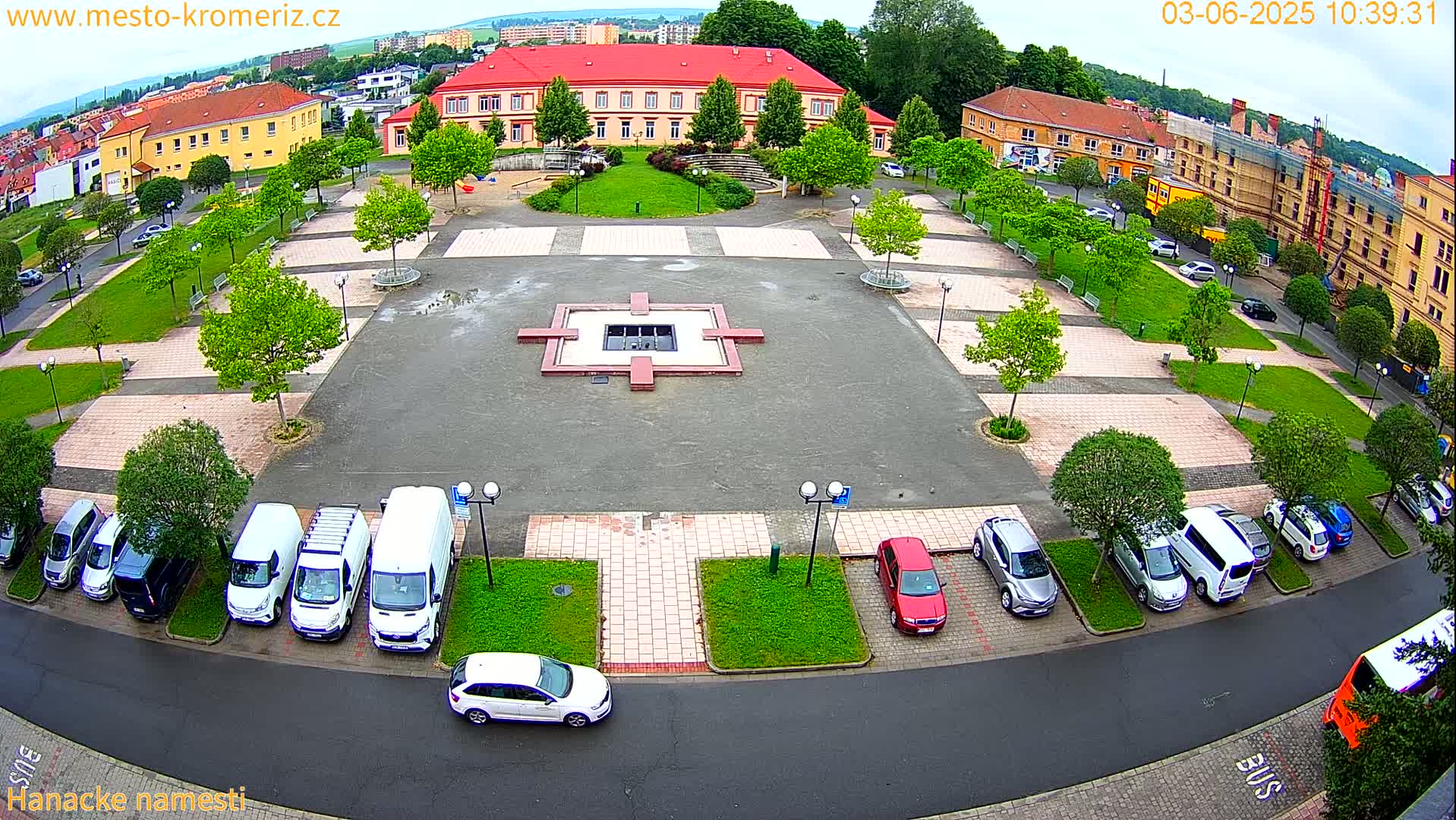 An aerial view of a town square with a central fountain, surrounded by parked cars and buildings, on a partly cloudy day.
