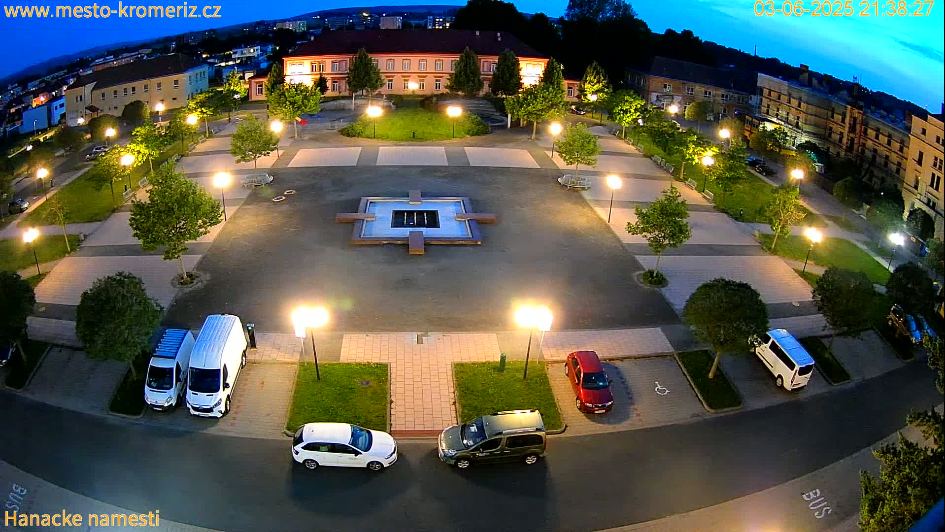 A town square at night, illuminated by streetlights, features a central fountain, surrounding green spaces, parked cars, and buildings in the background under a twilight sky.