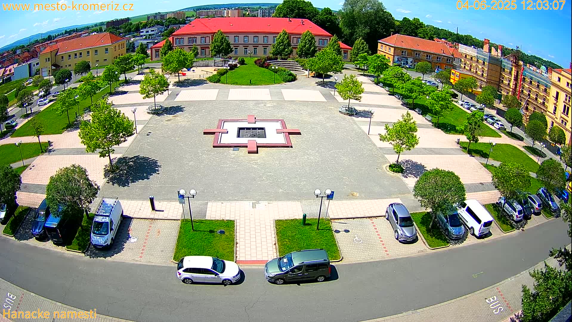 A town square with a central fountain, surrounded by buildings, trees, and parked cars under a sunny sky.