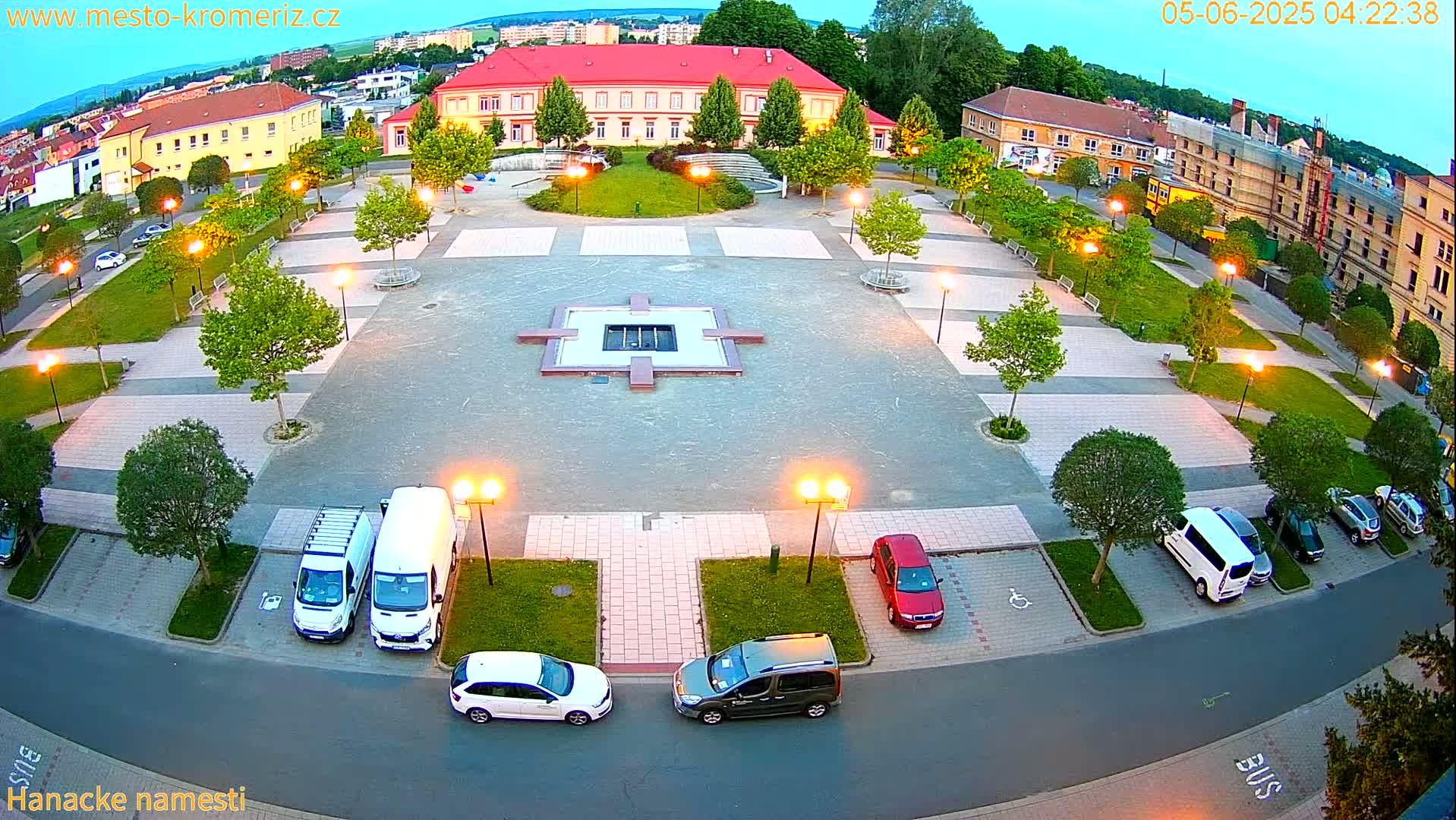 An illuminated town square at dusk features a central fountain, surrounding trees, parked cars, and a large building in the background.