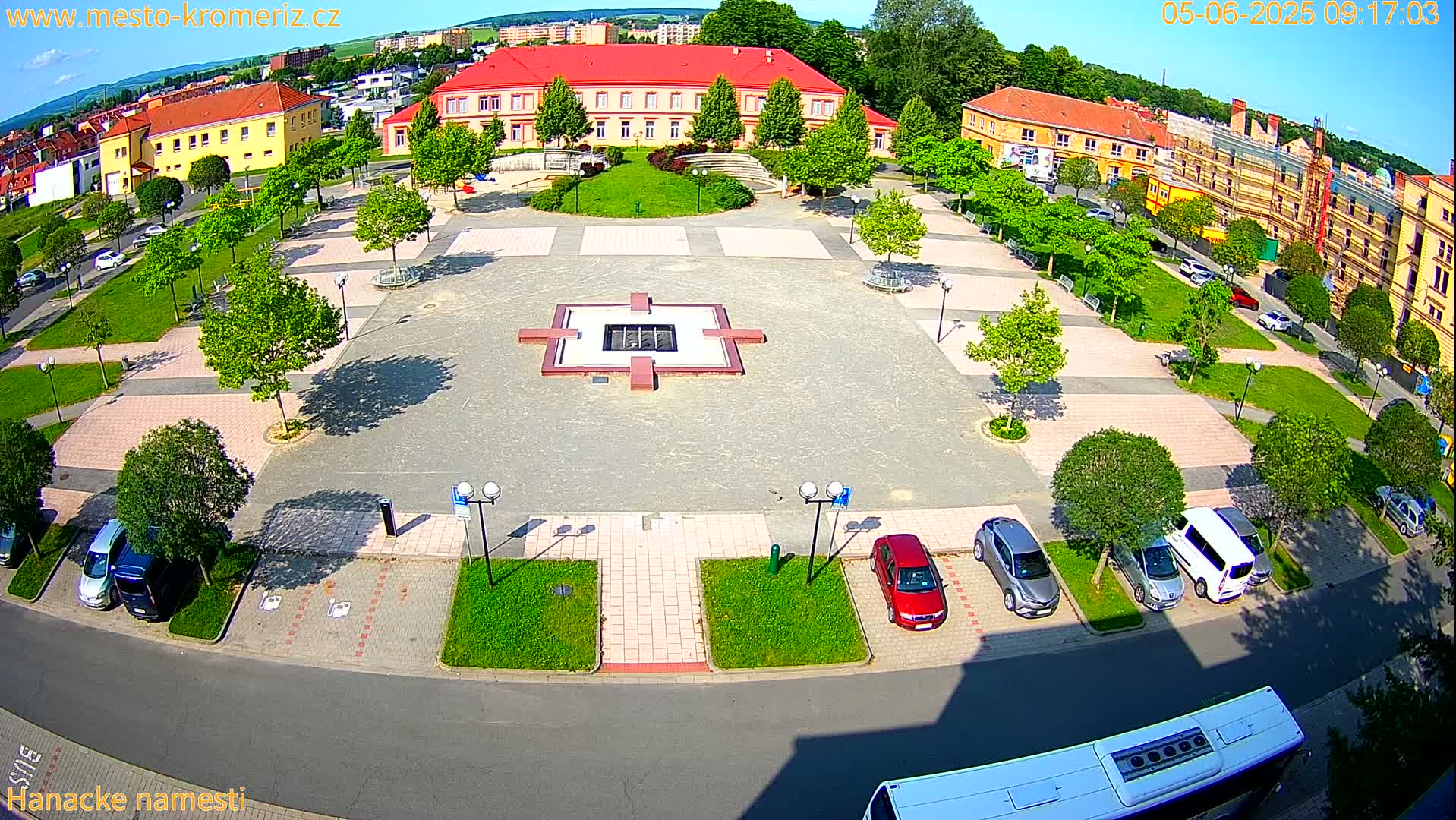 A sunny aerial view shows a town square with a central fountain, surrounded by parked cars, green lawns, and buildings.