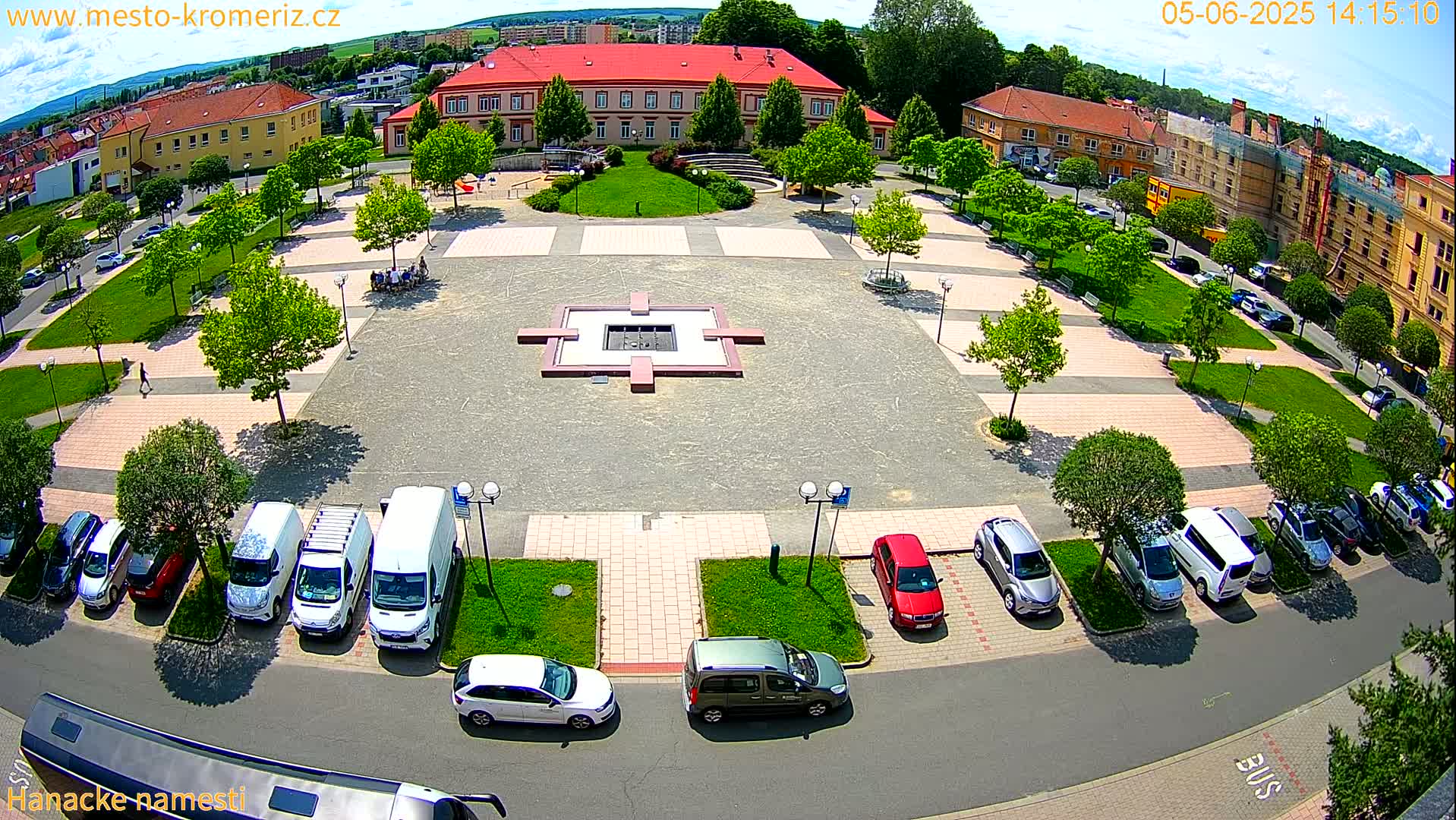 A high-angle view of a town square on a sunny day, showing a paved plaza with a central fountain, surrounded by buildings, parked cars, and green spaces.