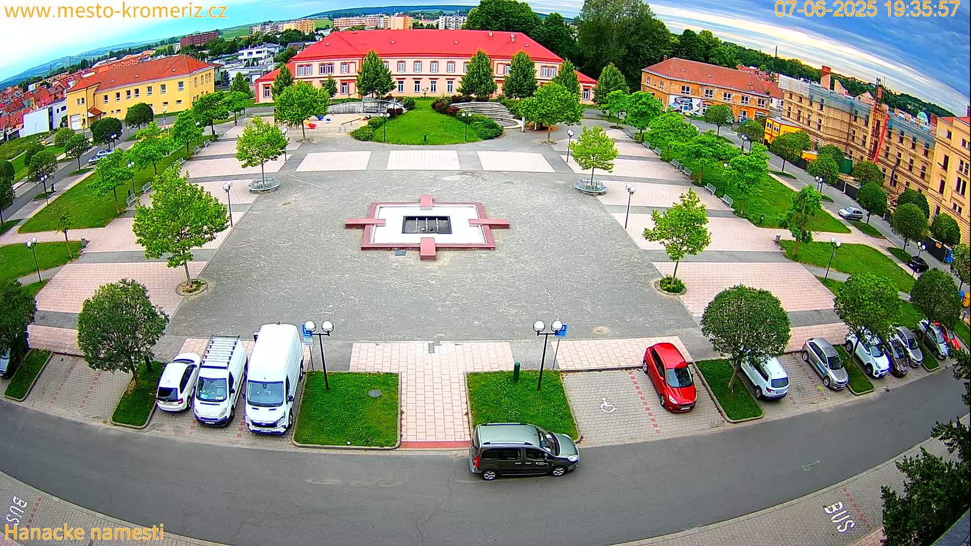An aerial view of a town square shows a paved plaza with a central fountain, surrounded by green lawns, trees, and parked cars under a partly cloudy sky.