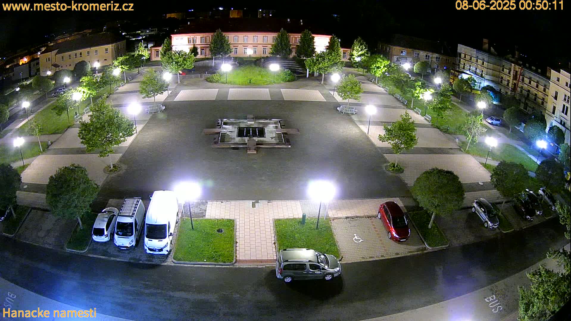 A nighttime aerial view of a town square with a fountain, several cars parked, and trees and lampposts lining the paved area.