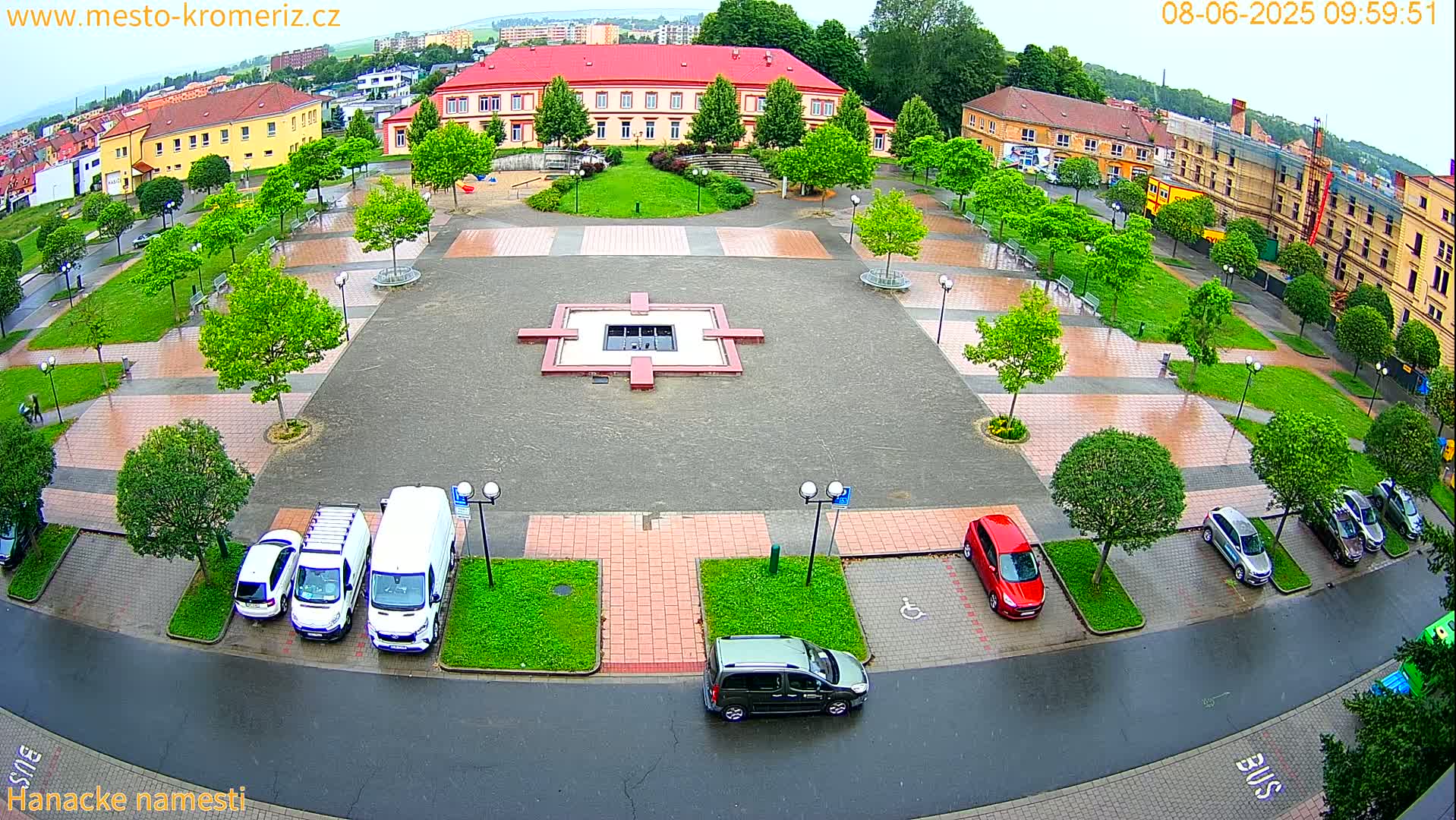 A wet town square with a central fountain, surrounded by parked cars and buildings, under an overcast sky.
