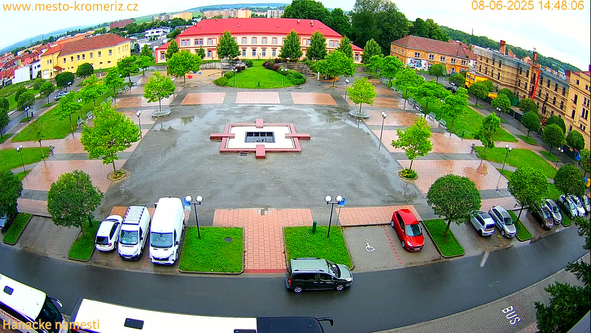 A wet town square with a fountain in the center, surrounded by parked cars, green spaces, and buildings, under an overcast sky.