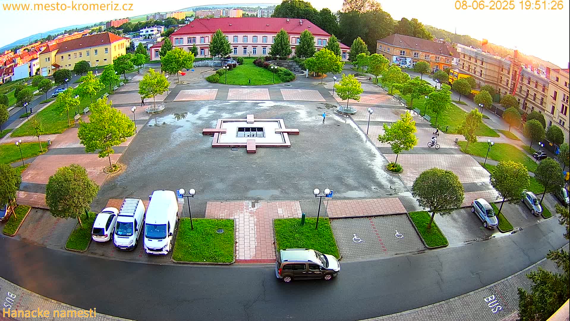An aerial view of a town square with a fountain, wet pavement, and several parked cars, surrounded by buildings and lush green trees under a partly cloudy evening sky.