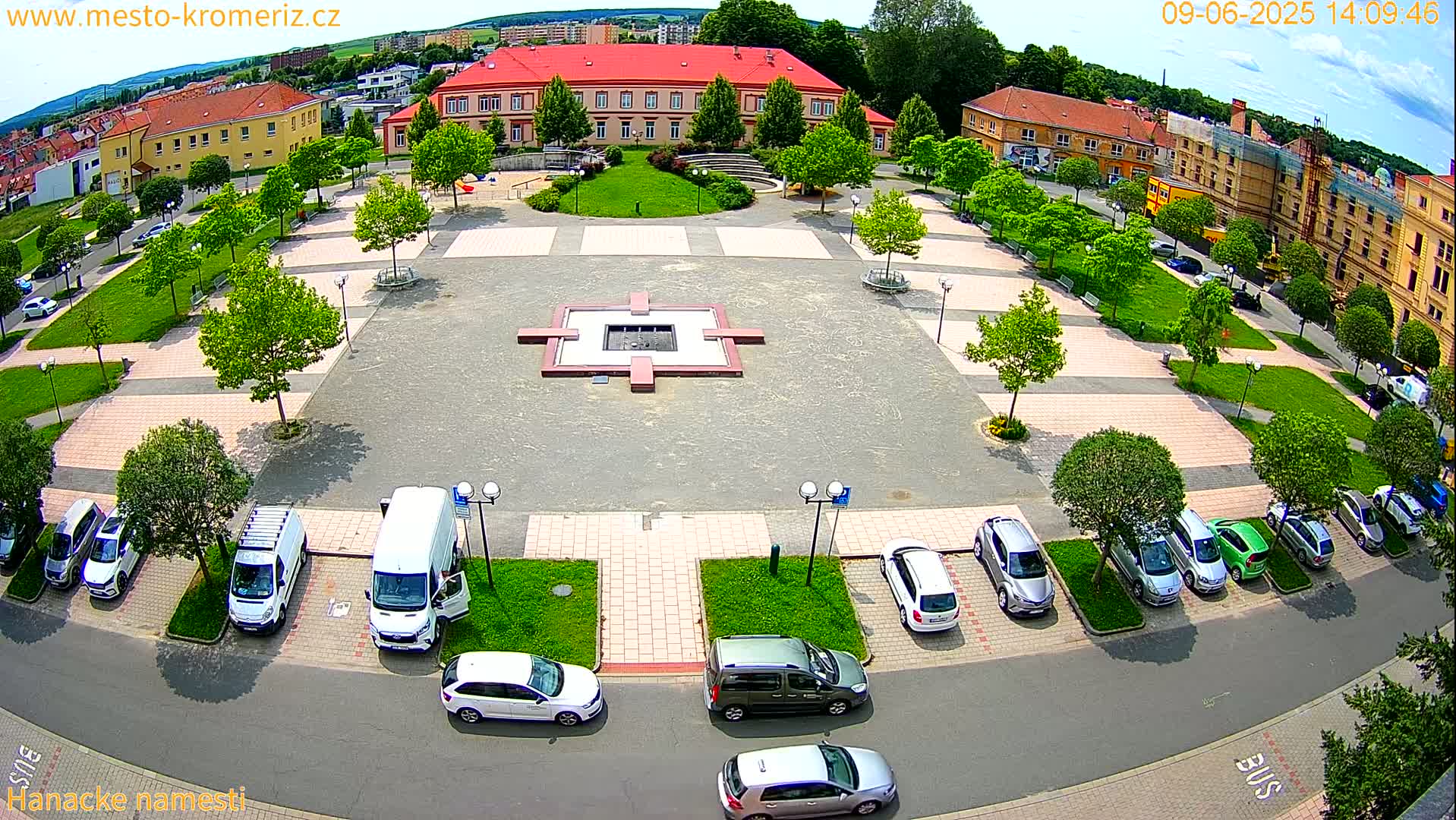 An aerial view of a town square on a sunny day, showing a central plaza with a small fountain, surrounded by buildings and cars parked along the edges.