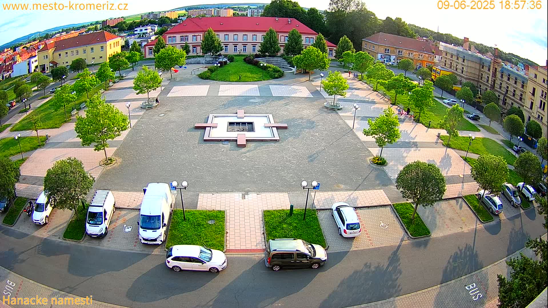 An aerial view of a town square with a central fountain, surrounded by buildings, trees, and parked cars on a sunny day.