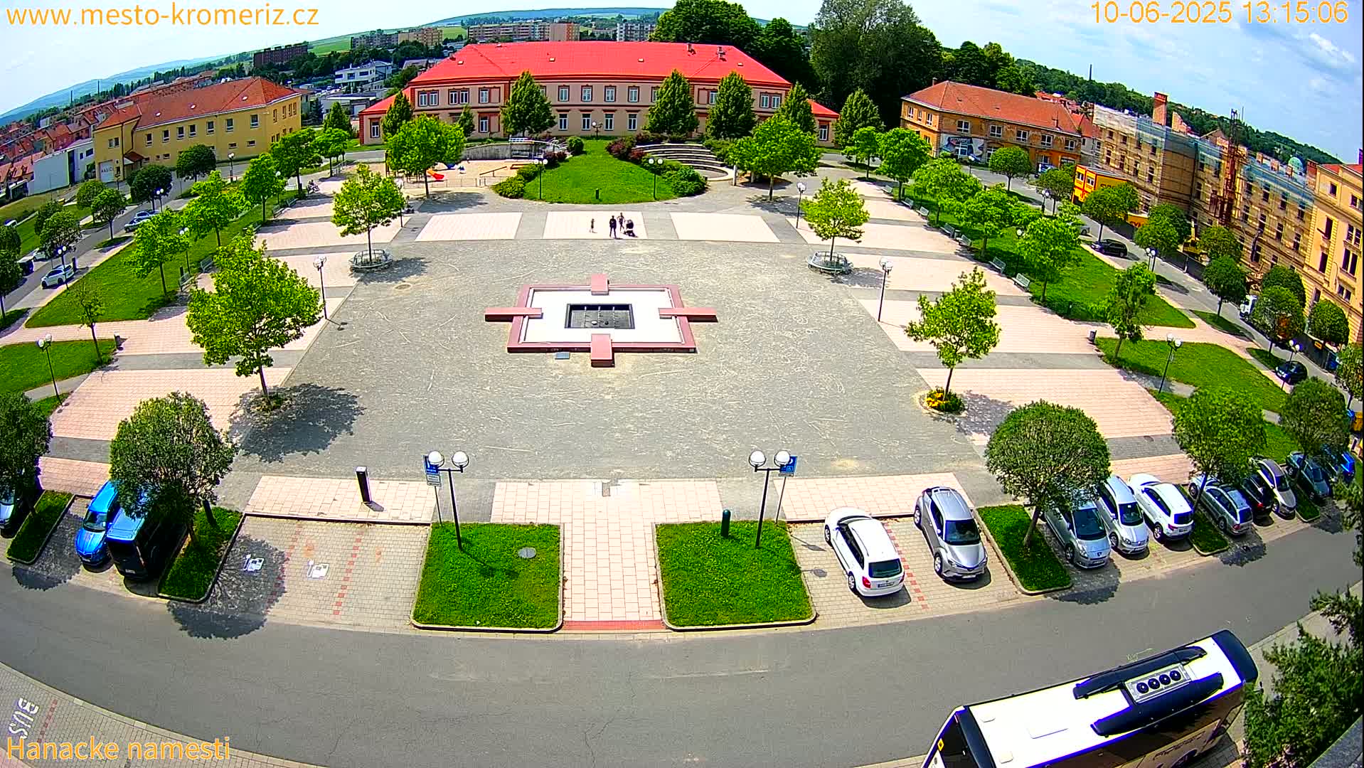 A town square with a central fountain, surrounded by buildings, green spaces, and parked cars under a sunny sky.