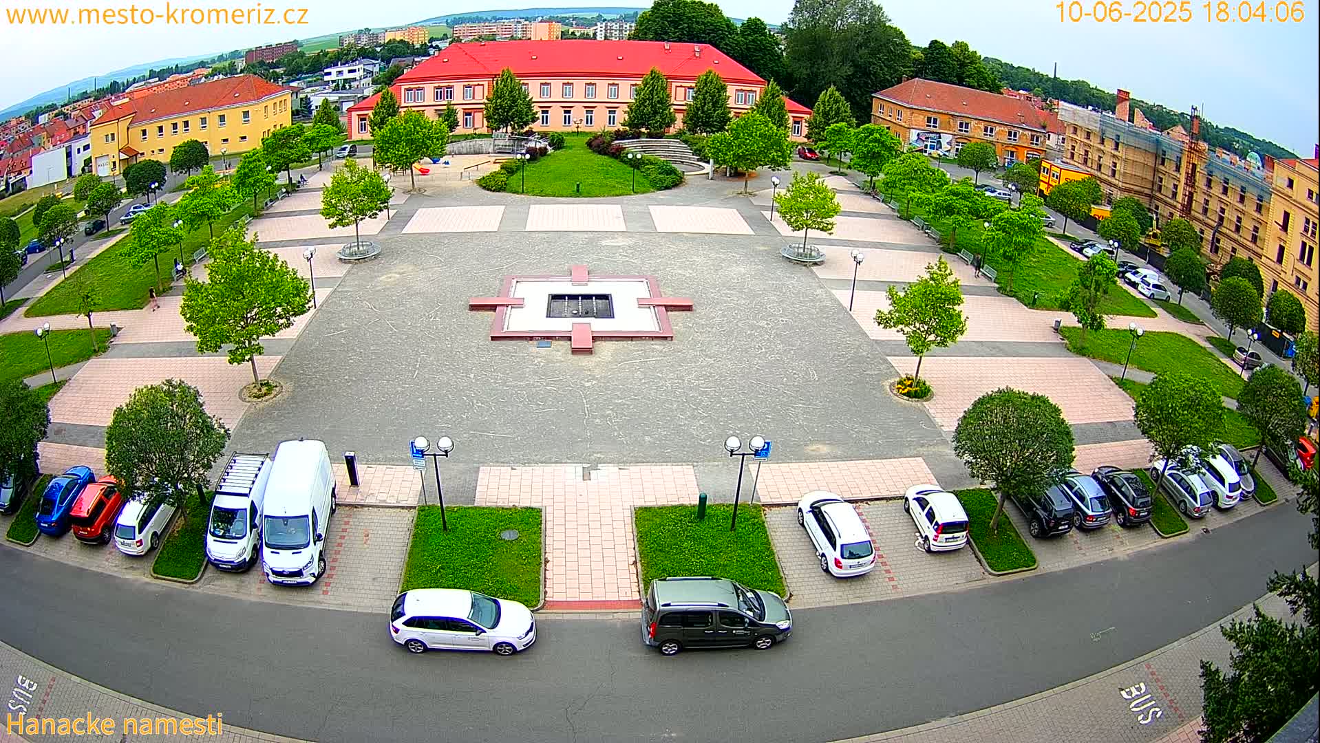 An aerial view of a town square with a central fountain, surrounded by parked cars, green lawns, and buildings under a partly cloudy sky.
