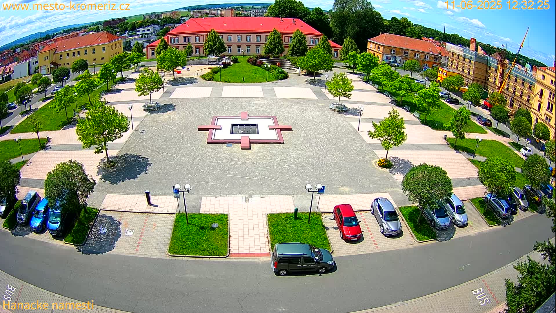 A sunny aerial view shows a town square with a central fountain, surrounded by parked cars, green spaces, and buildings.
