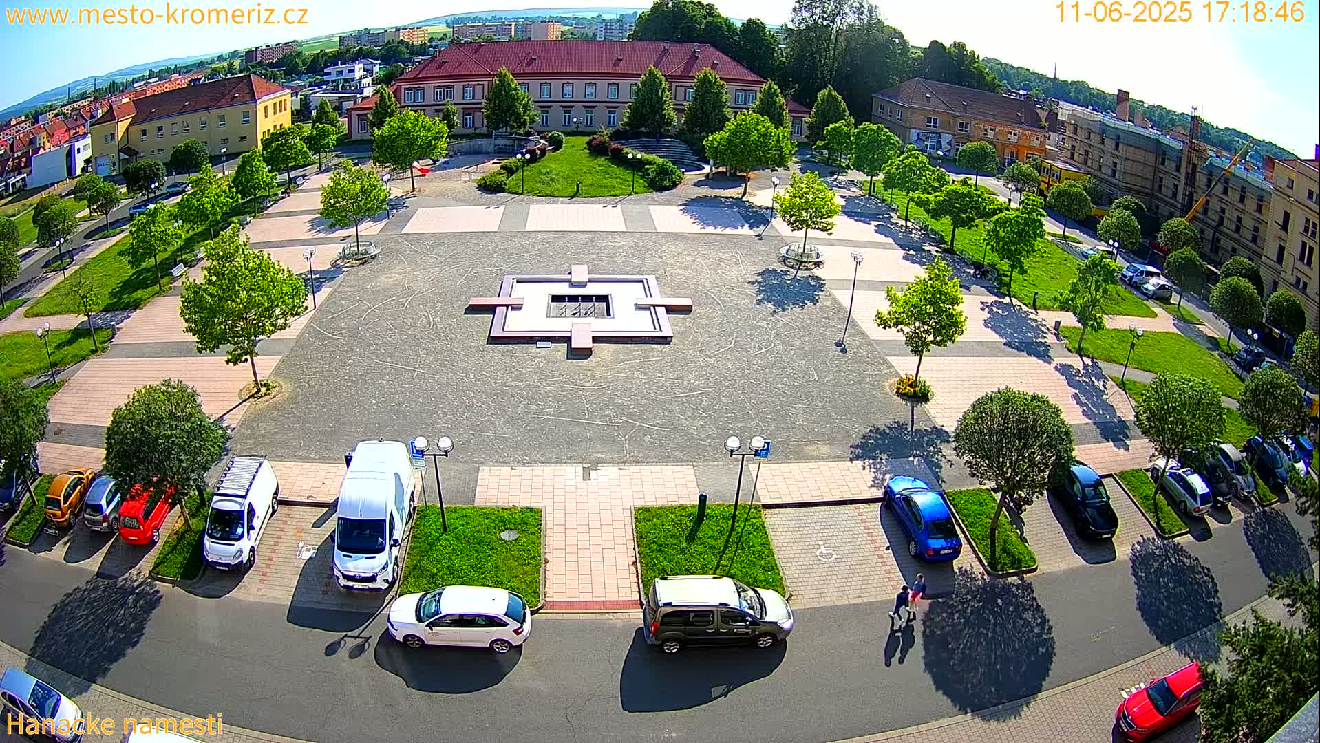 An aerial view of a town square with a central fountain, surrounded by parked cars and buildings, on a sunny day.