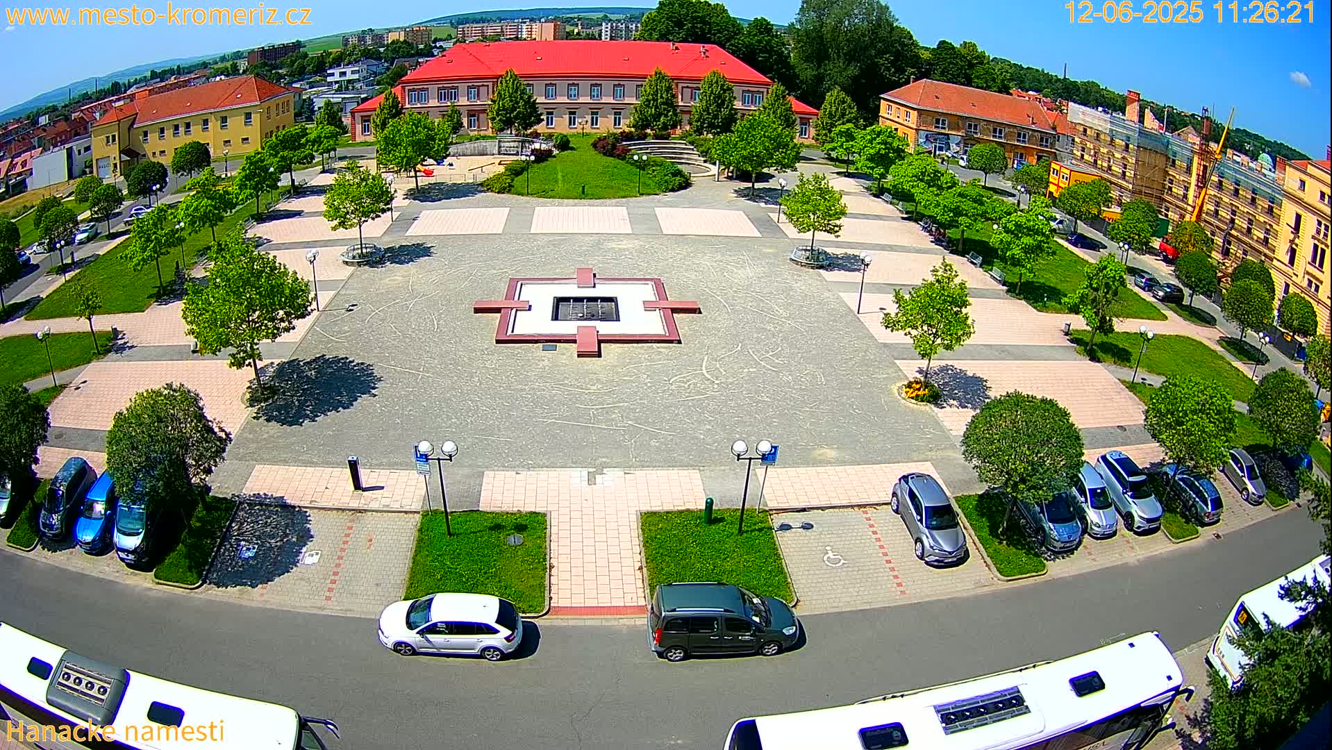 A large town square with a central fountain, surrounded by buildings, green spaces, and parked cars under a sunny sky.