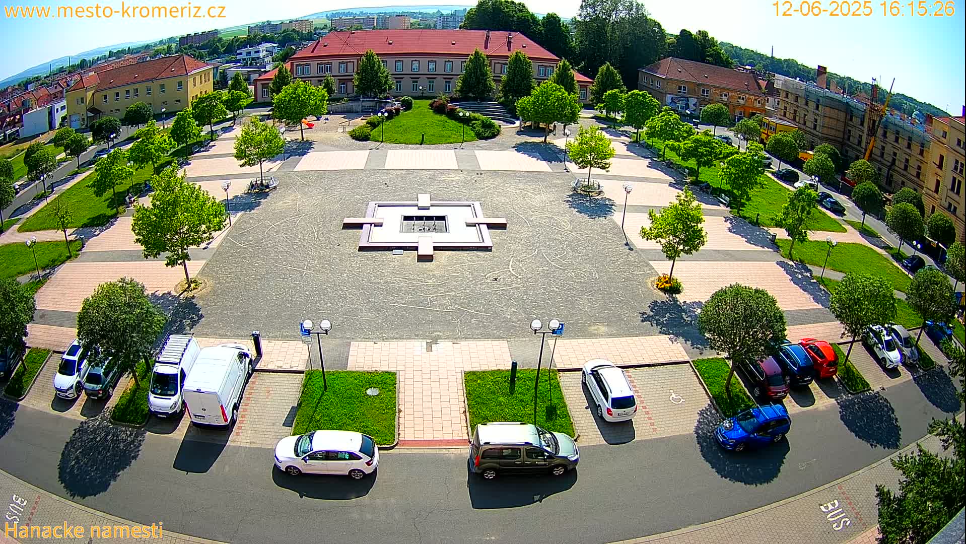 An aerial view of a town square with a fountain, surrounded by trees, buildings, and parked cars on a sunny day.
