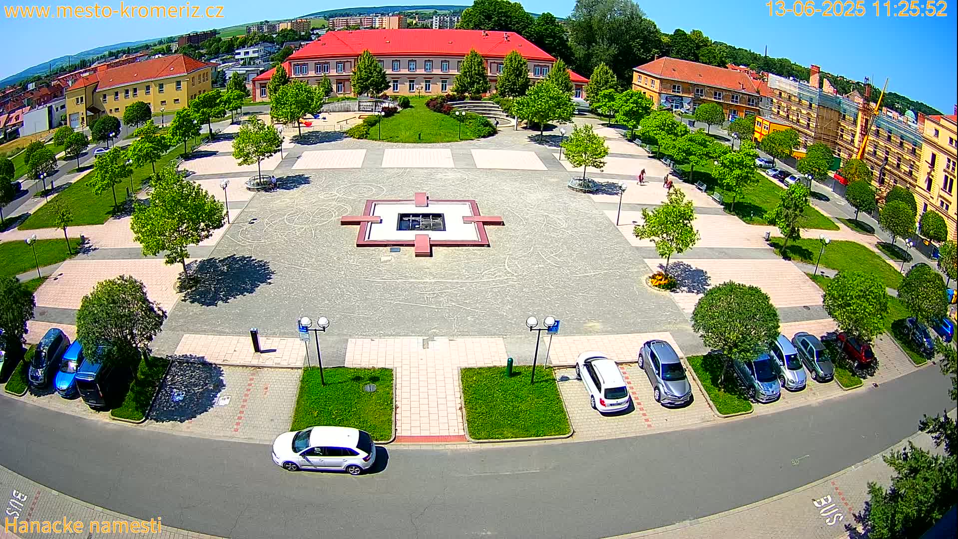 An aerial view of a town square on a sunny day shows a central fountain, surrounding green spaces with trees, and parked cars lining the edges.