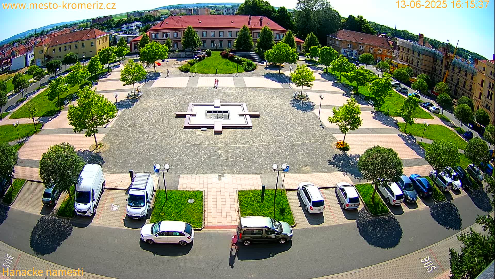 An aerial view of a town square on a sunny day shows a central fountain, surrounding green spaces, parked cars, and a large building in the background.