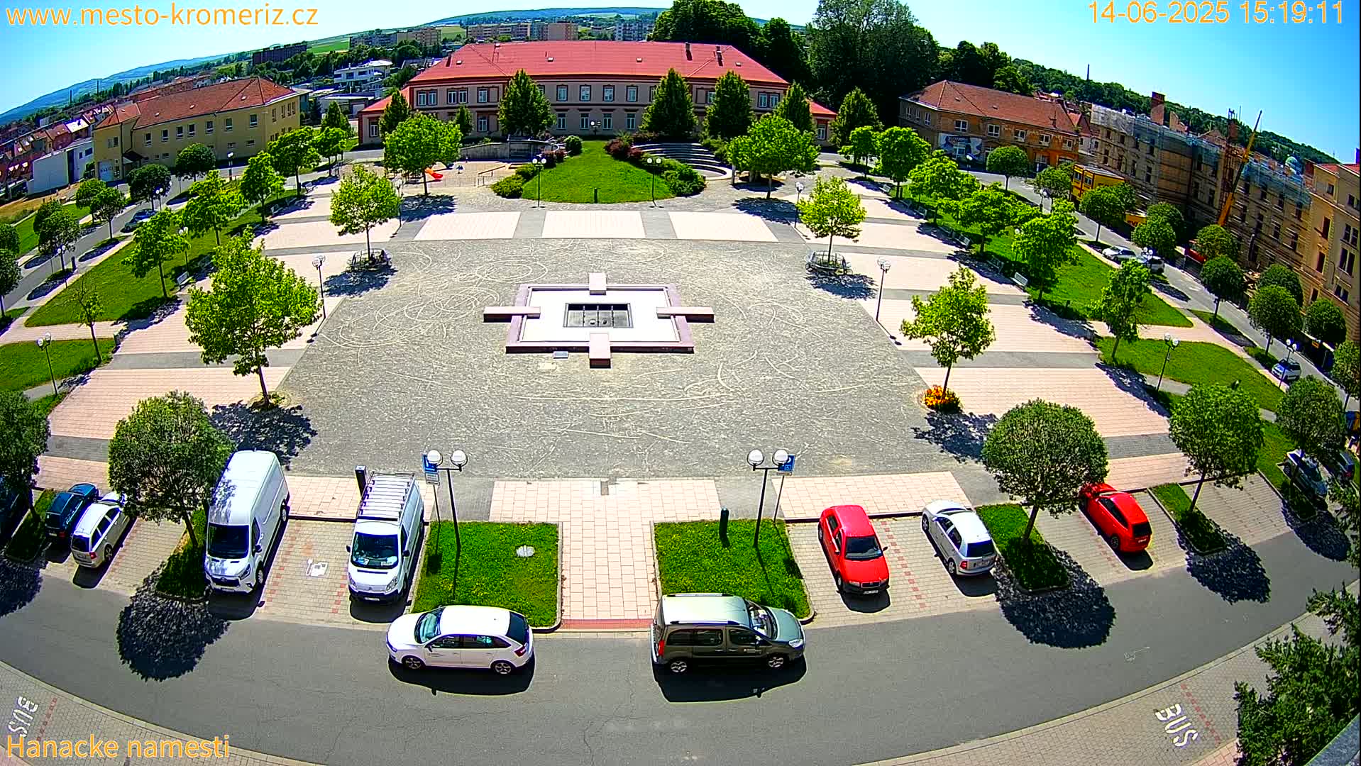 An aerial view of a town square on a sunny day shows a central fountain, surrounding green spaces, parked cars, and buildings in the background.