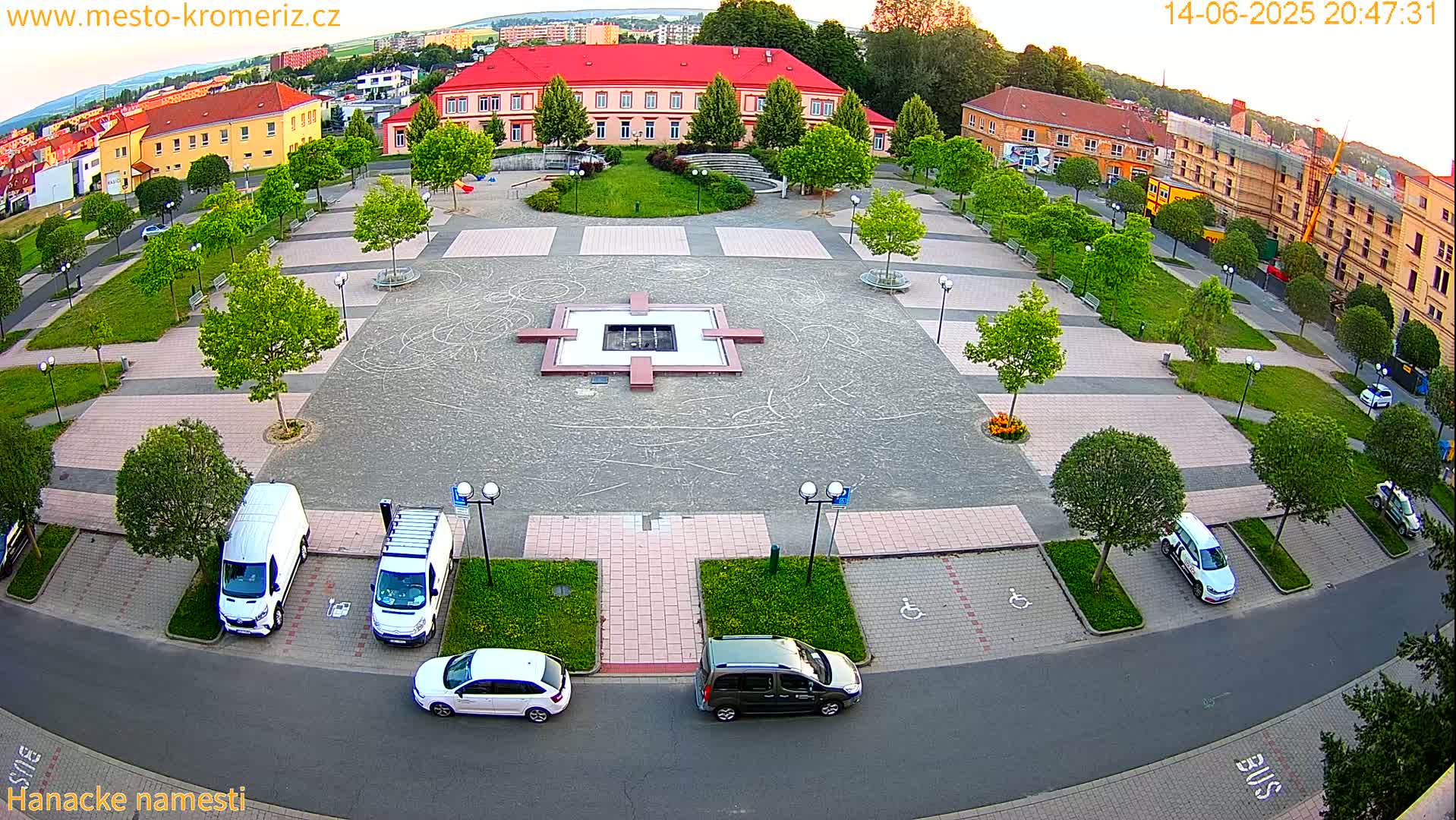 An aerial view of a town square on a sunny day shows a central fountain, surrounding green spaces, parked cars, and buildings in the background.