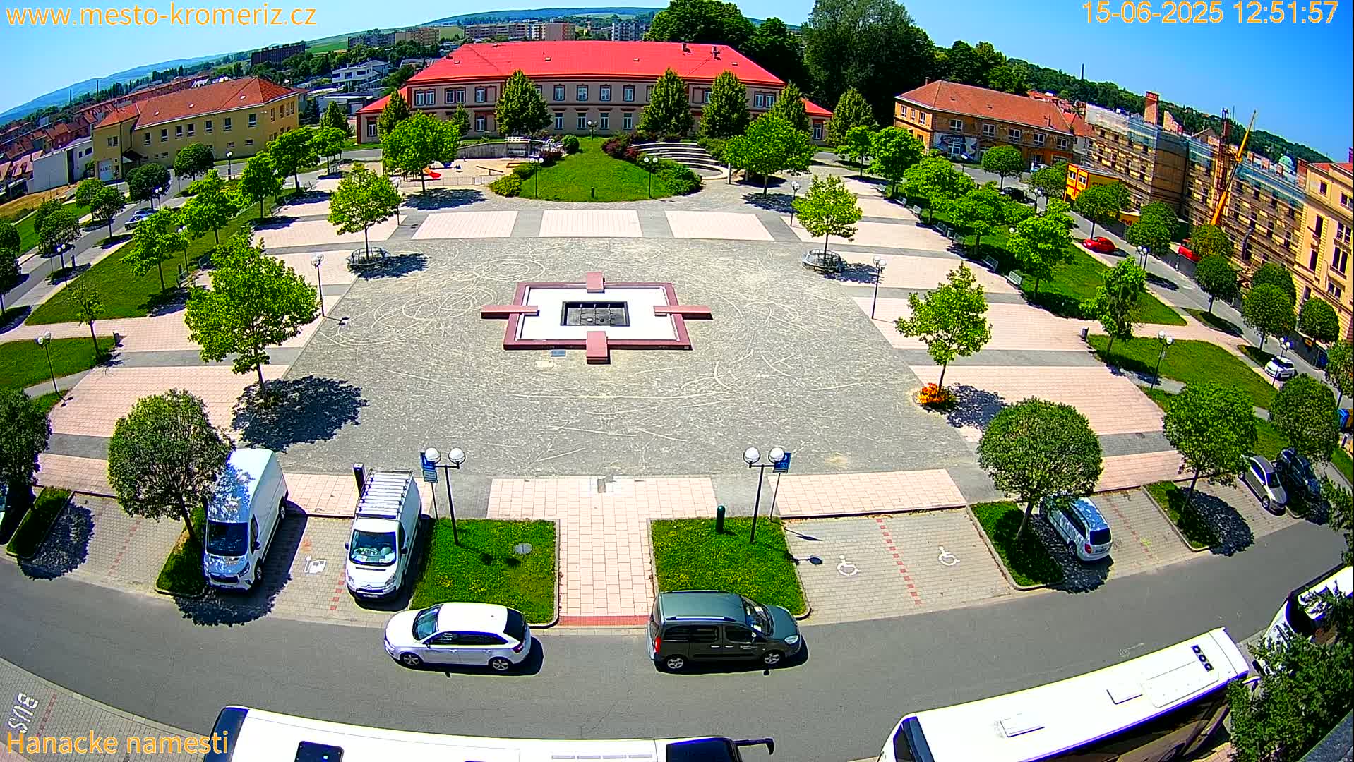 An aerial view of a town square on a sunny day, showing a paved area with a central fountain, surrounding landscaping, parked cars, and buildings.