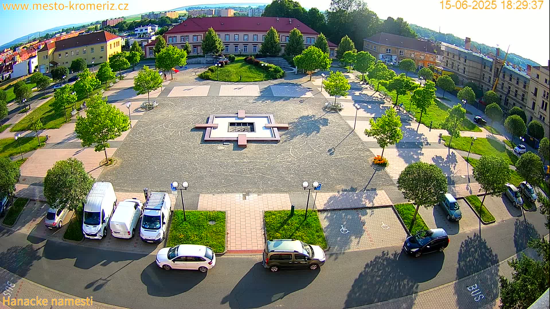 An aerial view of a town square with a central fountain, surrounded by parked cars and lush green trees under a sunny sky.